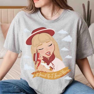 May include: A heather grey t-shirt featuring a graphic illustration of a blonde character wearing a red hat, holding a smaller doll, set against a sky with white clouds. A gold banner at the bottom displays the text I Knew It, I Knew You.