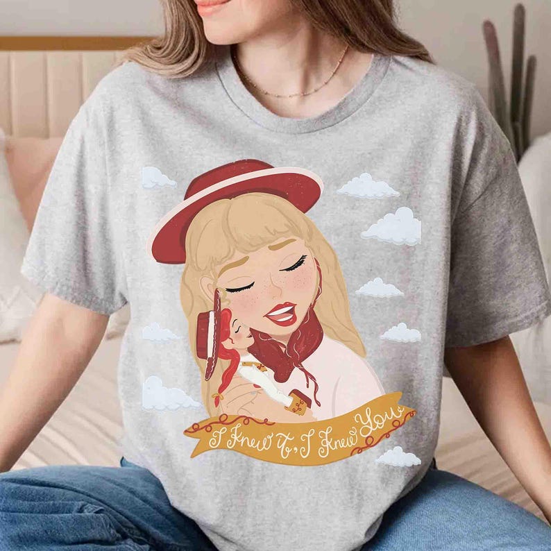 May include: A heather grey t-shirt featuring a graphic illustration of a blonde character wearing a red hat, holding a smaller doll, set against a sky with white clouds. A gold banner at the bottom displays the text I Knew It, I Knew You.