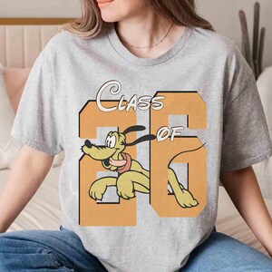 May include: A light gray t-shirt featuring the text "Class of 26" in a retro font, with a cartoon image of Pluto the dog. The dog is yellow with a red collar and is positioned within the number "26".