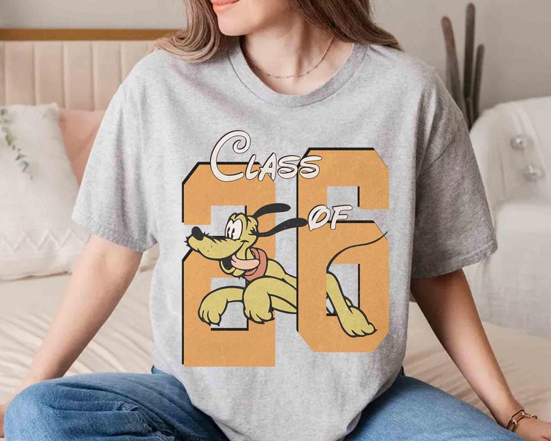 May include: A light gray t-shirt featuring the text "Class of 26" in a retro font, with a cartoon image of Pluto the dog. The dog is yellow with a red collar and is positioned within the number "26".
