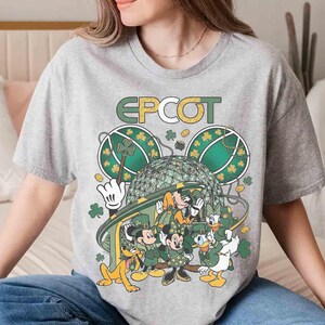 May include: A gray t-shirt featuring the word "EPCOT" in gold lettering. The shirt has a graphic of Disney characters dressed in green, with shamrocks and the EPCOT ball. The characters include Mickey, Minnie, Donald, Goofy, and Pluto.