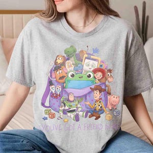 May include: A heather gray t-shirt featuring a graphic print of a purple backpack filled with various animated toy characters, including a cowboy, a space ranger, a dinosaur, and a pig, with the text "YOU'VE GOT A FRIEND IN ME" printed below.
