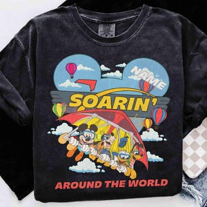 May include: Dark gray long-sleeve shirt featuring a colorful graphic of Mickey Mouse, Goofy, and Pluto hang gliding. The design includes hot air balloons, clouds, and the words "SOARIN'" and "AROUND THE WORLD".