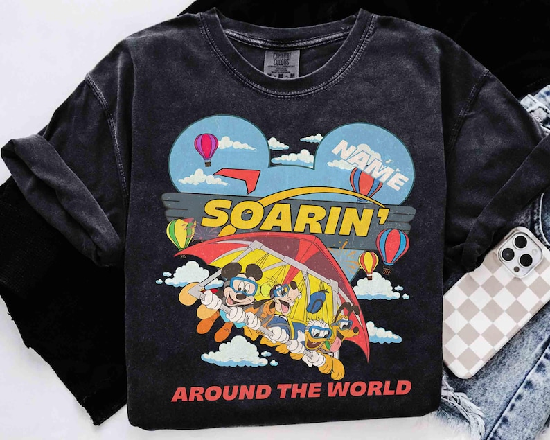 May include: Dark gray long-sleeve shirt featuring a colorful graphic of Mickey Mouse, Goofy, and Pluto hang gliding. The design includes hot air balloons, clouds, and the words "SOARIN'" and "AROUND THE WORLD".