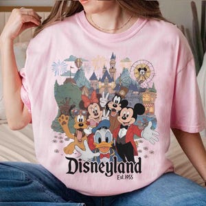 May include: Pink t-shirt featuring a Disneyland graphic with Mickey Mouse, Minnie Mouse, Donald Duck, Goofy, and other characters. The graphic includes a castle, fireworks, and a Ferris wheel. The text "Disneyland Est. 1955" is also visible.
