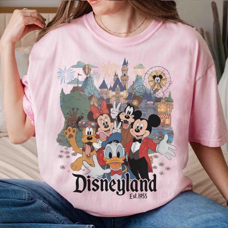 May include: Pink t-shirt featuring a Disneyland graphic with Mickey Mouse, Minnie Mouse, Donald Duck, Goofy, and other characters. The graphic includes a castle, fireworks, and a Ferris wheel. The text "Disneyland Est. 1955" is also visible.
