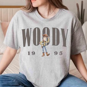 May include: A light gray t-shirt featuring the word "WOODY" in large, dark gray letters, with the numbers "1995" below. A cartoon image of Woody from Toy Story is in the center. The shirt is a casual, short-sleeved style.