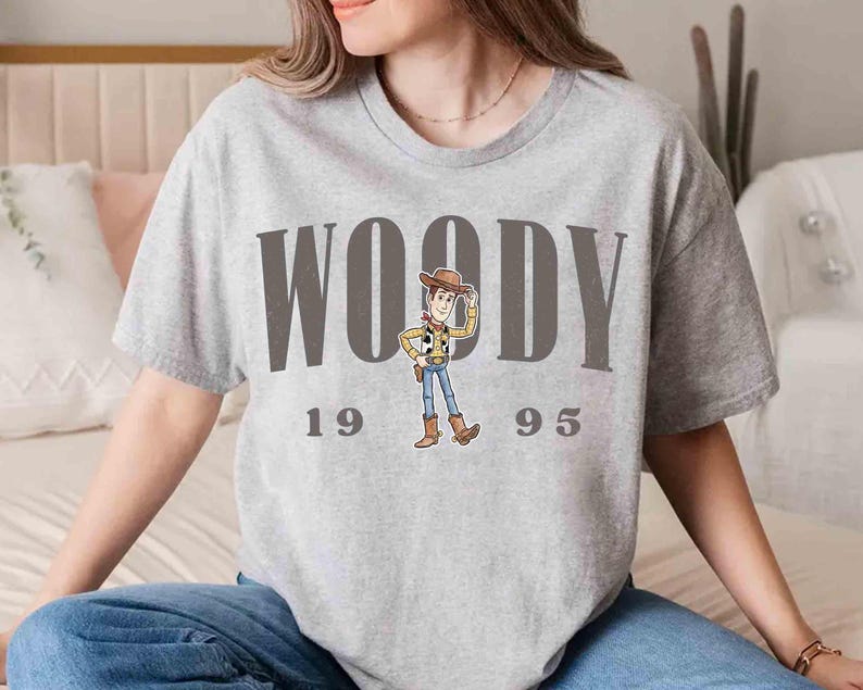 May include: A light gray t-shirt featuring the word "WOODY" in large, dark gray letters, with the numbers "1995" below. A cartoon image of Woody from Toy Story is in the center. The shirt is a casual, short-sleeved style.