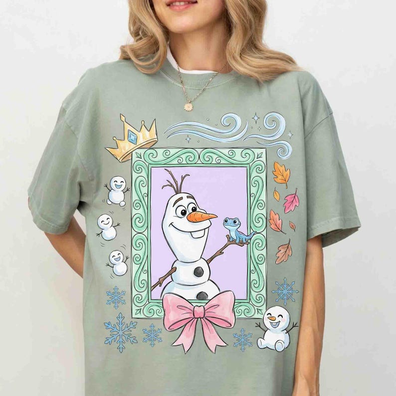 May include: A sage green t-shirt featuring a graphic print of a snowman and a small blue lizard inside a decorative rectangular frame. The design includes a gold crown, a large pink bow, falling autumn leaves, snowflakes, and small snowmen.
