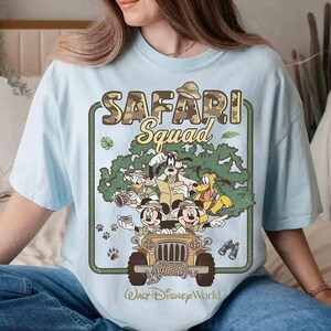 May include: Pale blue t-shirt with a Disney "SAFARI Squad" graphic. The design features Mickey Mouse, Donald Duck, Goofy, and Pluto in a jeep, with the text "Walt Disney World" below. The graphic has a camouflage and green tree design.