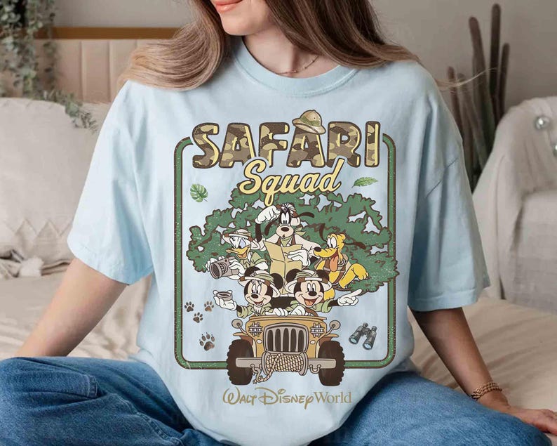 May include: Pale blue t-shirt with a Disney "SAFARI Squad" graphic. The design features Mickey Mouse, Donald Duck, Goofy, and Pluto in a jeep, with the text "Walt Disney World" below. The graphic has a camouflage and green tree design.