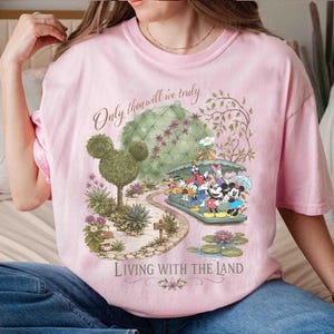 May include: Pink t-shirt featuring a whimsical illustration of Disney characters on a boat ride through a garden. The shirt includes the text "Only then will we truly" and "Living with the Land."