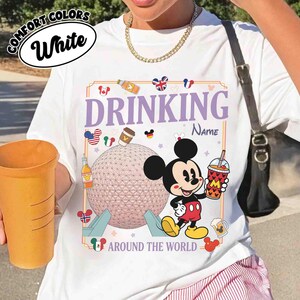 Puede incluir: Camiseta blanca con un gr&aacute;fico de Mickey Mouse sosteniendo una bebida, el texto "DRINKING AROUND THE WORLD" e iconos de pa&iacute;ses. La camiseta tambi&eacute;n tiene el texto "COMFORT COLORS White".