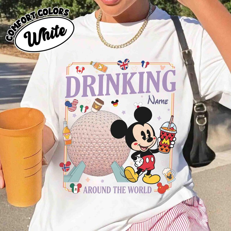 Puede incluir: Camiseta blanca con un gr&aacute;fico de Mickey Mouse sosteniendo una bebida, el texto "DRINKING AROUND THE WORLD" e iconos de pa&iacute;ses. La camiseta tambi&eacute;n tiene el texto "COMFORT COLORS White".