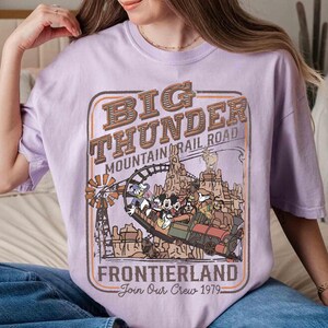 May include: A light purple t-shirt featuring a graphic of the Big Thunder Mountain Railroad ride. The design includes the text "Big Thunder Mountain Railroad Frontierland Join Our Crew 1979" and cartoon characters.