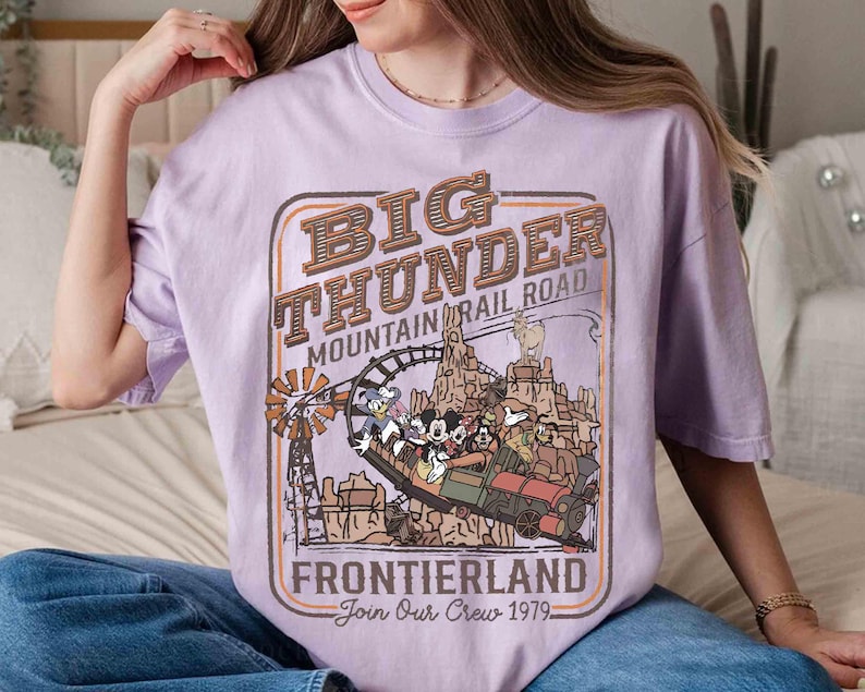 May include: A light purple t-shirt featuring a graphic of the Big Thunder Mountain Railroad ride. The design includes the text "Big Thunder Mountain Railroad Frontierland Join Our Crew 1979" and cartoon characters.