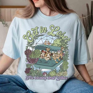 May include: Light blue t-shirt with a graphic featuring cartoon characters in a ride vehicle with the text "Get in Loser" and "We're learning about Lettuce". The design includes green and purple foliage.
