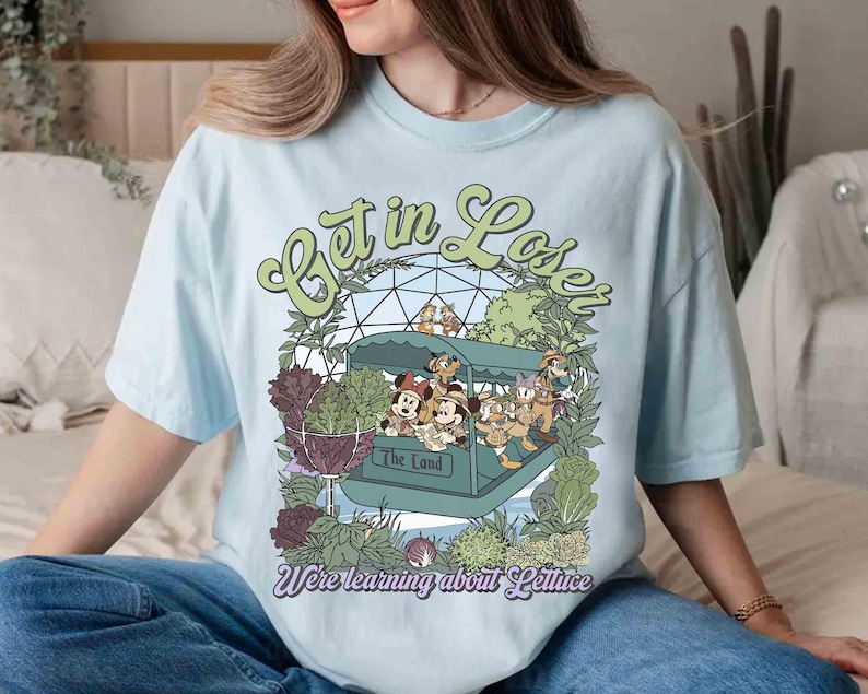 May include: Light blue t-shirt with a graphic featuring cartoon characters in a ride vehicle with the text "Get in Loser" and "We're learning about Lettuce". The design includes green and purple foliage.