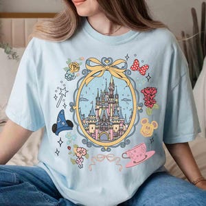 May include: Light blue t-shirt featuring a graphic of Cinderella's castle, Mickey Mouse, and other Disney characters. The graphic is surrounded by a floral design and a ribbon.