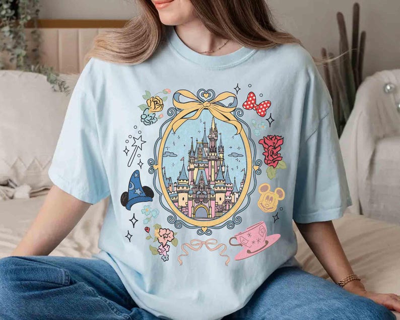 May include: Light blue t-shirt featuring a graphic of Cinderella's castle, Mickey Mouse, and other Disney characters. The graphic is surrounded by a floral design and a ribbon.