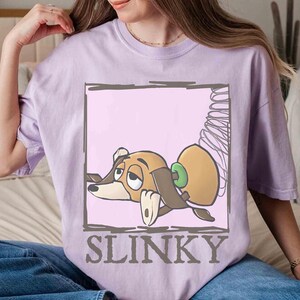 May include: A light purple t-shirt featuring a cartoon image of Slinky Dog from Toy Story. The graphic shows Slinky in a rectangular frame with the word "SLINKY" below the image. The shirt is made of a soft material.