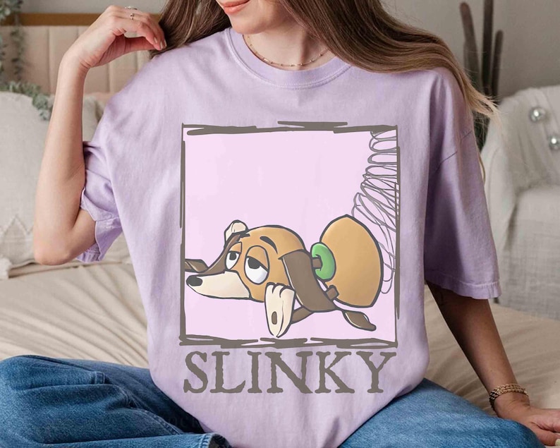 May include: A light purple t-shirt featuring a cartoon image of Slinky Dog from Toy Story. The graphic shows Slinky in a rectangular frame with the word "SLINKY" below the image. The shirt is made of a soft material.