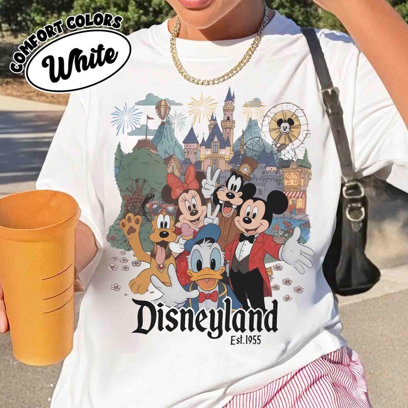 May include: White t-shirt featuring a colorful Disneyland graphic with Mickey Mouse, Minnie Mouse, Donald Duck, Goofy, and other characters. The shirt has the text "Disneyland Est.1955" in a black script font.