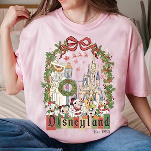 Op de afbeelding: Roze T-shirt met een Disneyland kerstontwerp. De afbeelding toont een kasteel, Mickey Mouse, Donald Duck en Goofy, omringd door feestelijke versieringen en de tekst "Disneyland Est. 1955".