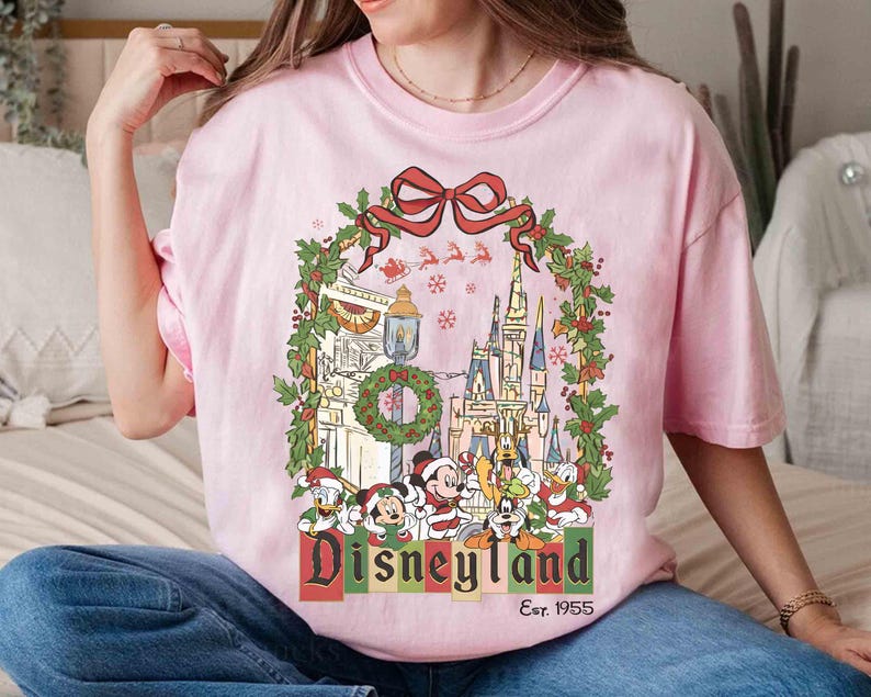 Op de afbeelding: Roze T-shirt met een Disneyland kerstontwerp. De afbeelding toont een kasteel, Mickey Mouse, Donald Duck en Goofy, omringd door feestelijke versieringen en de tekst "Disneyland Est. 1955".
