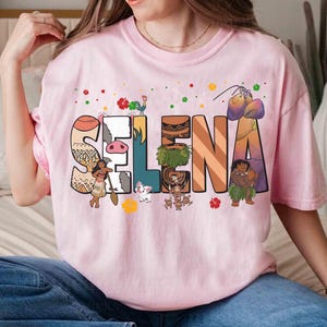 Puede incluir: Camiseta rosa claro con el nombre "Selena" en letras decorativas y coloridas. Cada letra est&aacute; dise&ntilde;ada con un patr&oacute;n y personaje diferente de una pel&iacute;cula de animaci&oacute;n popular. La camiseta es de estilo casual y c&oacute;modo.