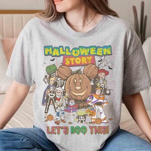 Puede incluir: Una camiseta gris con un gr&aacute;fico colorido de Halloween. El dise&ntilde;o presenta una calabaza sonriente con orejas de Mickey Mouse, rodeada de personajes de dibujos animados y el texto "HALLOWEEN STORY" y "LET'S BOO THIS!"