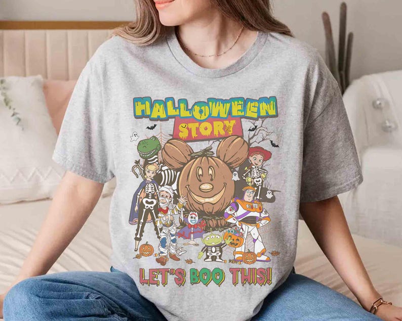 Puede incluir: Una camiseta gris con un gr&aacute;fico colorido de Halloween. El dise&ntilde;o presenta una calabaza sonriente con orejas de Mickey Mouse, rodeada de personajes de dibujos animados y el texto "HALLOWEEN STORY" y "LET'S BOO THIS!"