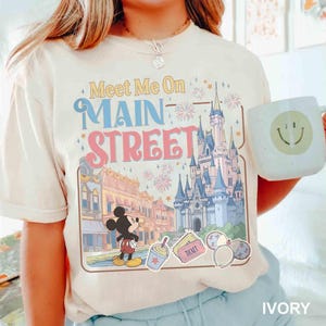 May include: Ivory-colored t-shirt featuring a colorful graphic of a castle, Main Street, and Mickey Mouse. The text "Meet Me On Main Street" is displayed above the graphic. A white mug with a smiley face is held in the image.