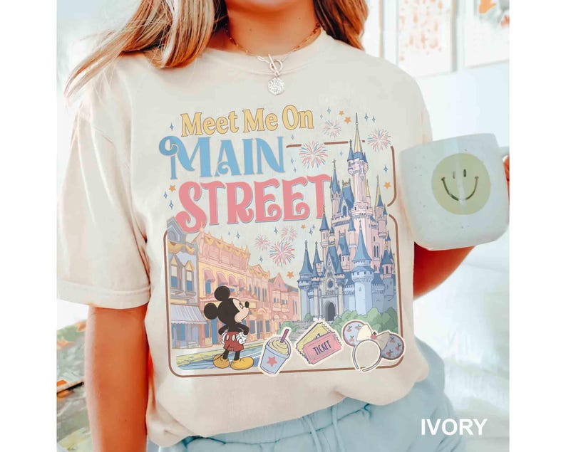 May include: Ivory-colored t-shirt featuring a colorful graphic of a castle, Main Street, and Mickey Mouse. The text "Meet Me On Main Street" is displayed above the graphic. A white mug with a smiley face is held in the image.