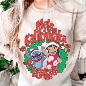 May include: Beige sweatshirt featuring the phrase "Mele Kalikimaka" in red script. The graphic includes cartoon characters Stitch and Lilo, surrounded by red flowers and green leaves, all wearing Santa hats. A festive holiday design.