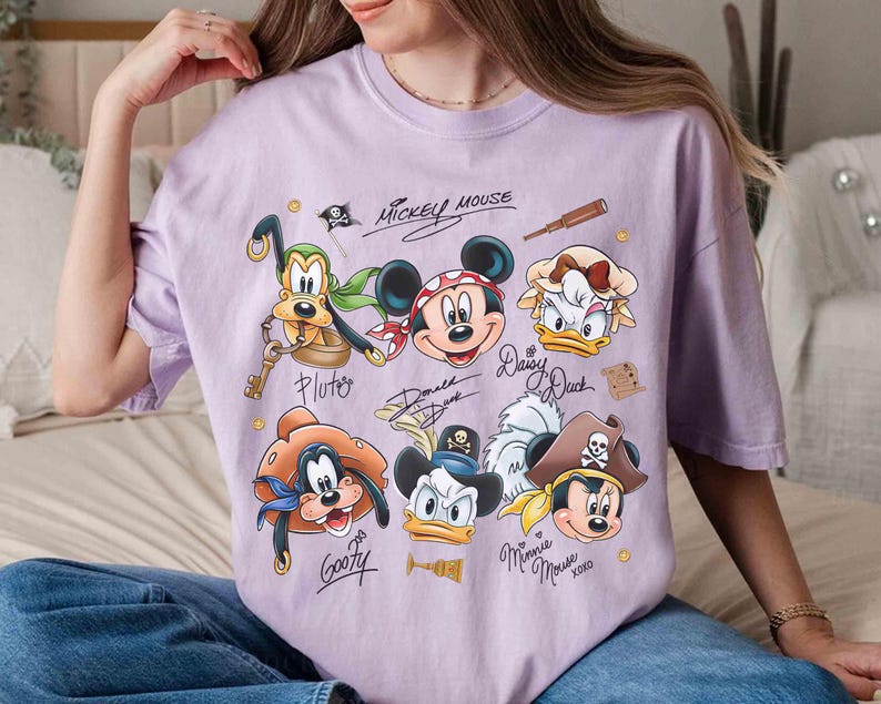 May include: A light purple t-shirt featuring a graphic of Mickey Mouse and friends in pirate attire. The design includes Pluto, Mickey, Daisy, Goofy, Donald, and Minnie Mouse, with their signatures. The words "Mickey Mouse" are also printed.
