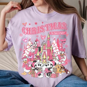 May include: A light purple t-shirt with a graphic featuring Mickey and Minnie Mouse, Donald Duck, Goofy, and other characters in front of a pink castle and Christmas trees. The text "CHRISTMAS ON MAIN STREET" is above the graphic.