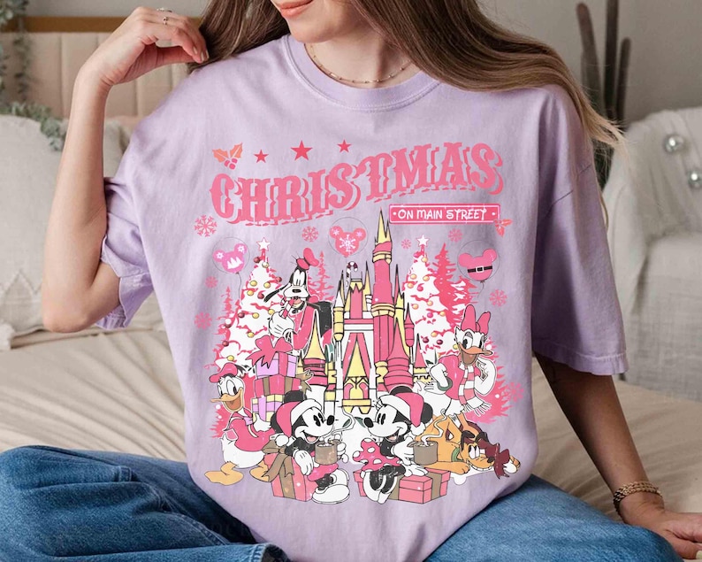 May include: A light purple t-shirt with a graphic featuring Mickey and Minnie Mouse, Donald Duck, Goofy, and other characters in front of a pink castle and Christmas trees. The text "CHRISTMAS ON MAIN STREET" is above the graphic.