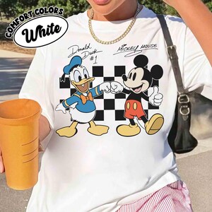 May include: White t-shirt featuring Donald Duck and Mickey Mouse fist-bumping in front of a black and white checkered background. The shirt has the text "Donald Duck #1" and "Mickey Mouse" in script. The words "Comfort Colors" and "White" are in a white oval.