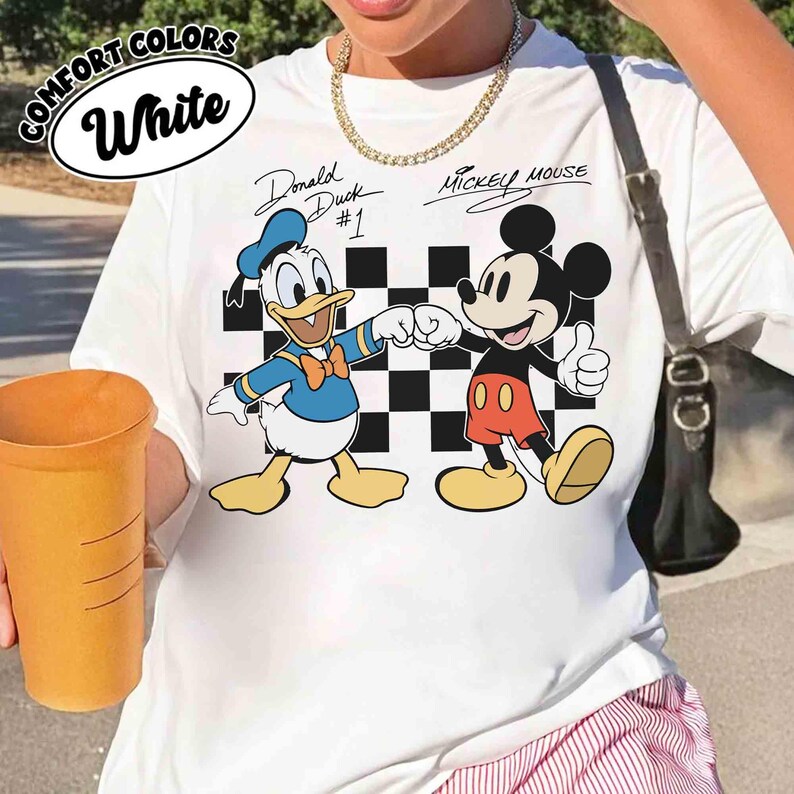 May include: White t-shirt featuring Donald Duck and Mickey Mouse fist-bumping in front of a black and white checkered background. The shirt has the text "Donald Duck #1" and "Mickey Mouse" in script. The words "Comfort Colors" and "White" are in a white oval.