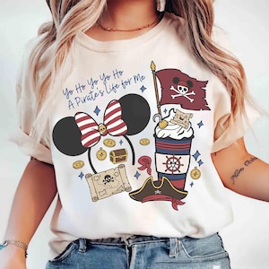 Könnte beinhalten: Cremefarbenes T-Shirt mit einem Piraten-Motiv. Das Design umfasst eine von Minnie Mouse inspirierte Schleife, eine Piratenflagge, eine Schatzkarte, Münzen und eine Kaffeetasse. Der Text lautet "Yo Ho Yo Ho A Pirate's Life for Me."