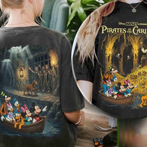 May include: Dark gray t-shirt featuring a colorful graphic of Disney characters in a boat, with a pirate-themed design. The back of the shirt shows a scene with a dog and prisoners behind bars. The text "Disney Mickey & Friends Pirates of the Caribbean" is printed on the shirt.