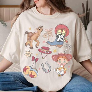May include: Cream-coloured t-shirt with cartoon illustrations of a cowboy theme. The design includes a horse, cowboy boots, a hat, a sheriff's badge, and a cowgirl character. The shirt has a relaxed fit.
