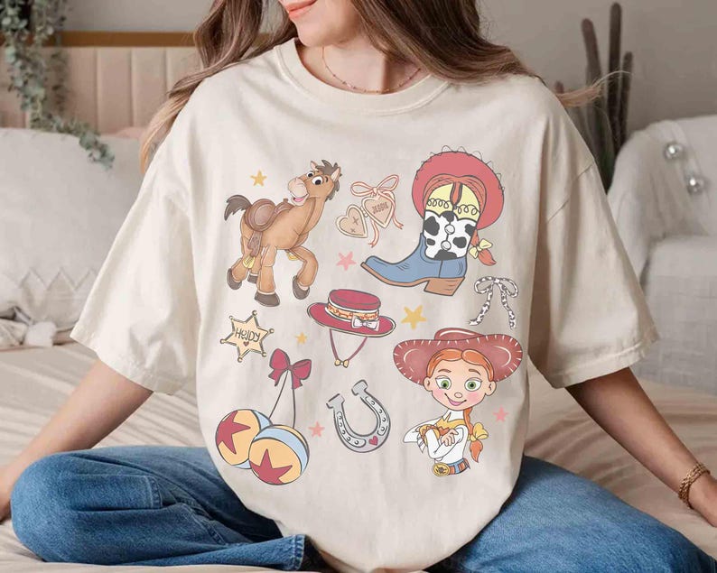 May include: Cream-coloured t-shirt with cartoon illustrations of a cowboy theme. The design includes a horse, cowboy boots, a hat, a sheriff's badge, and a cowgirl character. The shirt has a relaxed fit.