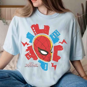 Puede incluir: Una camiseta azul claro con un gr&aacute;fico de la m&aacute;scara de Spider-Man en rojo y negro, rodeada de siluetas de edificios urbanos rojos y azules y formas de rayos, con el texto Spider-Man Brand New Day impreso debajo.