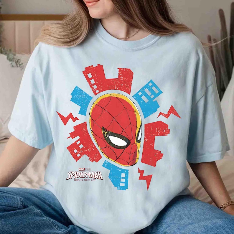 Puede incluir: Una camiseta azul claro con un gr&aacute;fico de la m&aacute;scara de Spider-Man en rojo y negro, rodeada de siluetas de edificios urbanos rojos y azules y formas de rayos, con el texto Spider-Man Brand New Day impreso debajo.