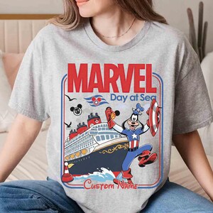 May include: A gray t-shirt featuring a graphic with the text "MARVEL Day at Sea." The design includes a cruise ship, the Marvel logo, and a cartoon character dressed as Captain America. The words "Custom Name" are also visible.
