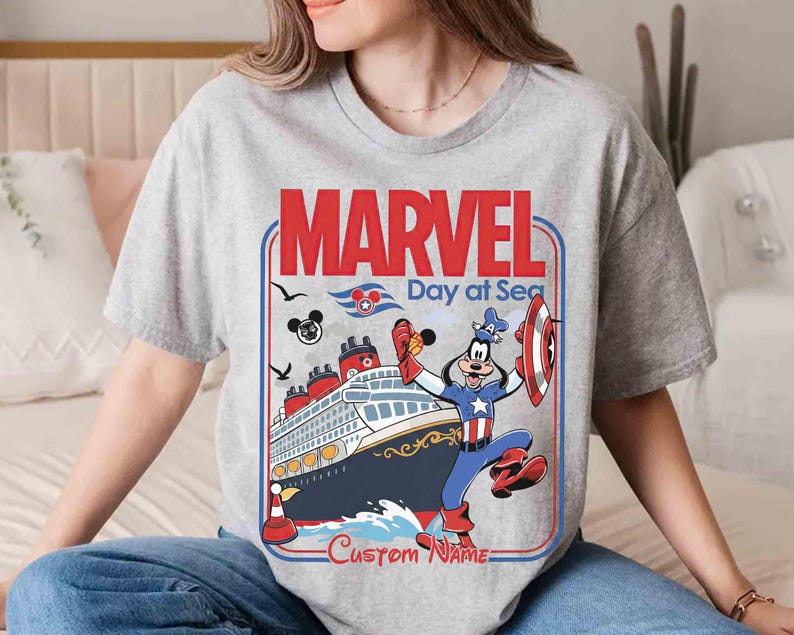 May include: A gray t-shirt featuring a graphic with the text "MARVEL Day at Sea." The design includes a cruise ship, the Marvel logo, and a cartoon character dressed as Captain America. The words "Custom Name" are also visible.