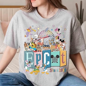 May include: A gray t-shirt featuring a colorful graphic of Disney characters and the text "EPCOT Since 1982." The design includes Mickey and Minnie Mouse, Donald Duck, and other characters, with the EPCOT sphere in the background.