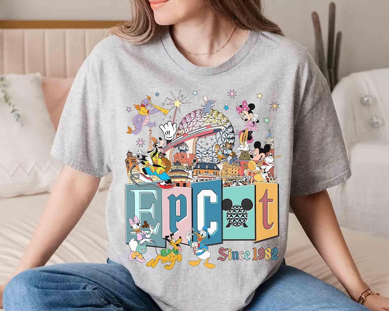 May include: A gray t-shirt featuring a colorful graphic of Disney characters and the text "EPCOT Since 1982." The design includes Mickey and Minnie Mouse, Donald Duck, and other characters, with the EPCOT sphere in the background.